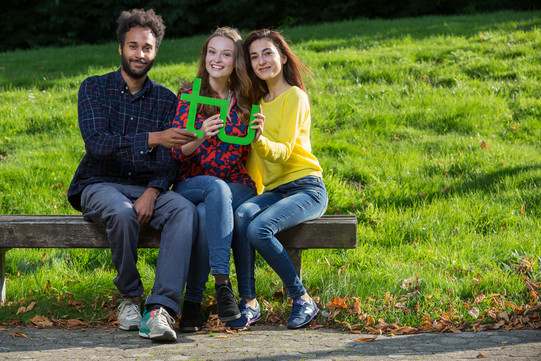 Drei Studierende sitzen auf einer Bank und halten gemeinsam ein grünes TU-Logo in den Händen.